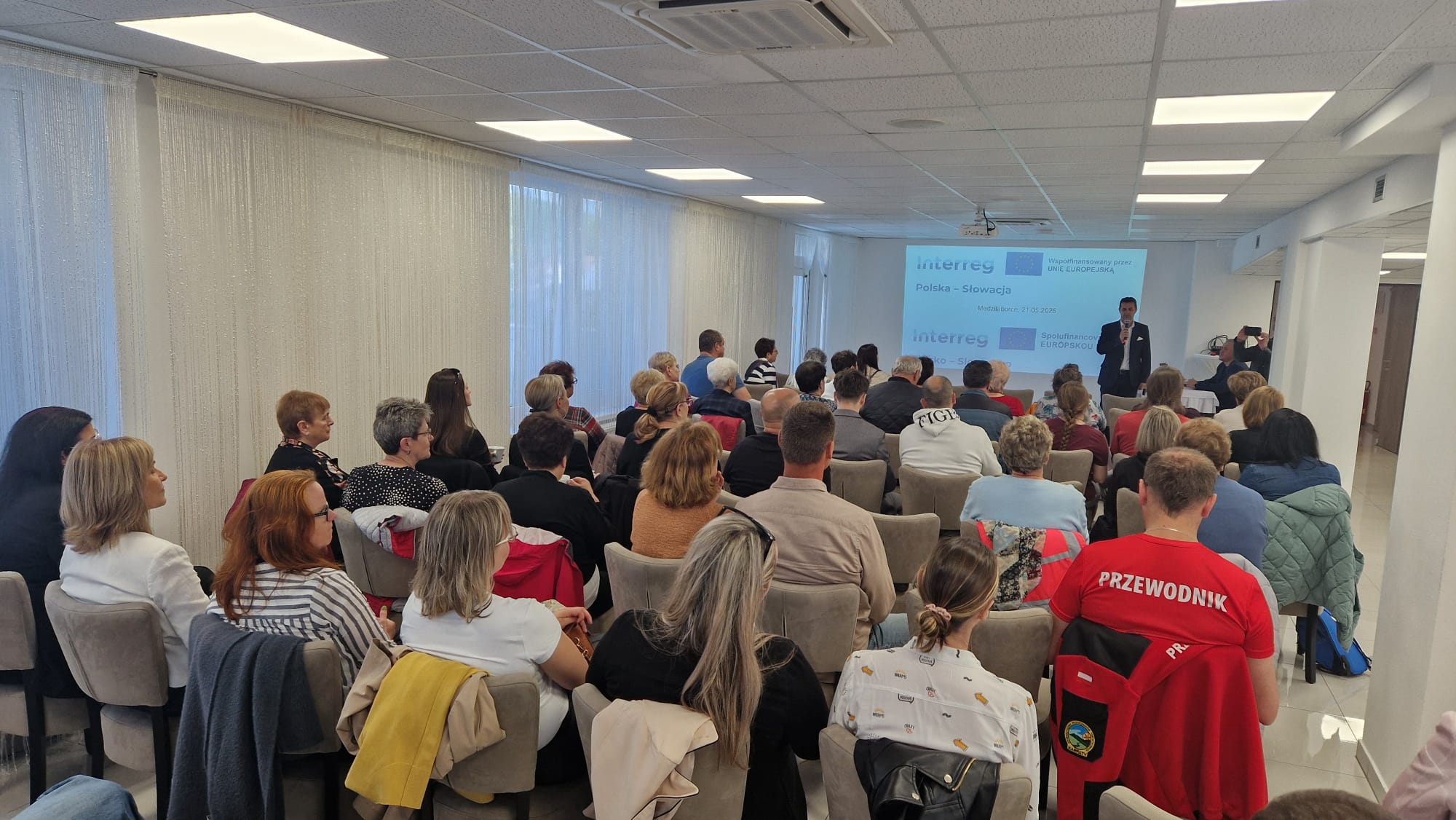 Sala konferencyjna, w kt&oacute;rej trwa prezentacja. Widzowie siedzą na krzesłach, a prelegent stoi przed ekranem z logiem Interreg.